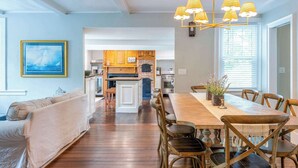 A spacious living room with a wooden floor, a dining area with a large table and chairs, and a kitchen in the background.
