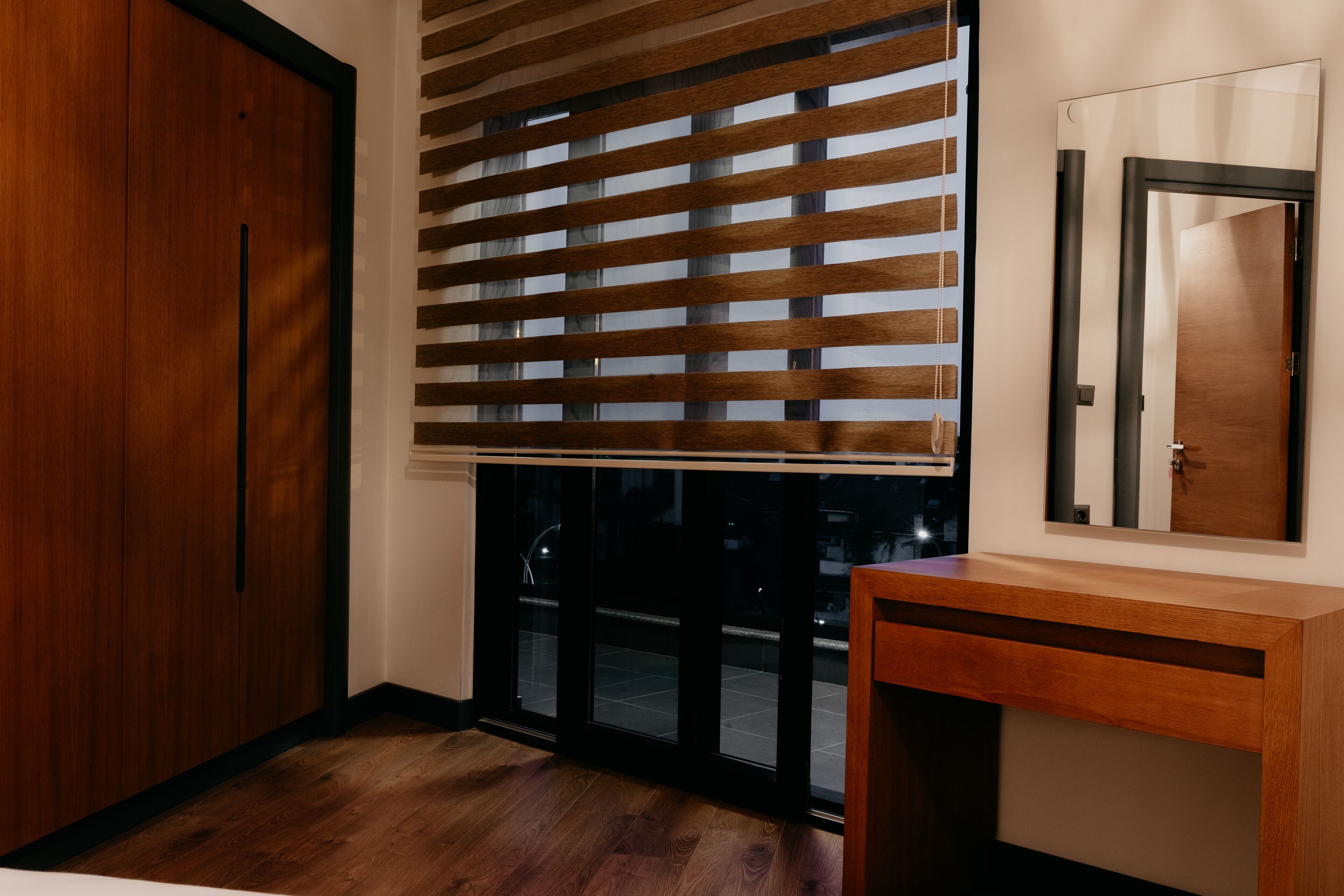 A room with a wooden wardrobe, a sliding glass door with blinds, a wooden console table, and a mirror on the wall.