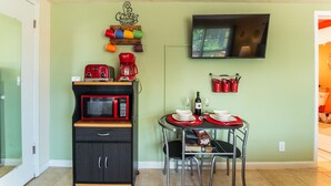 Une cuisine compacte équipée d’un micro-ondes, d’un grille-pain et d’une machine à café, le tout posé sur une étagère en bois, ainsi qu’une petite table à manger avec des chaises et un téléviseur fixé au mur.