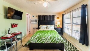 Une chambre avec un lit, un téléviseur fixé au mur, une fenêtre avec des rideaux, un ventilateur de plafond et une petite table avec des verres à vin et des bouteilles.