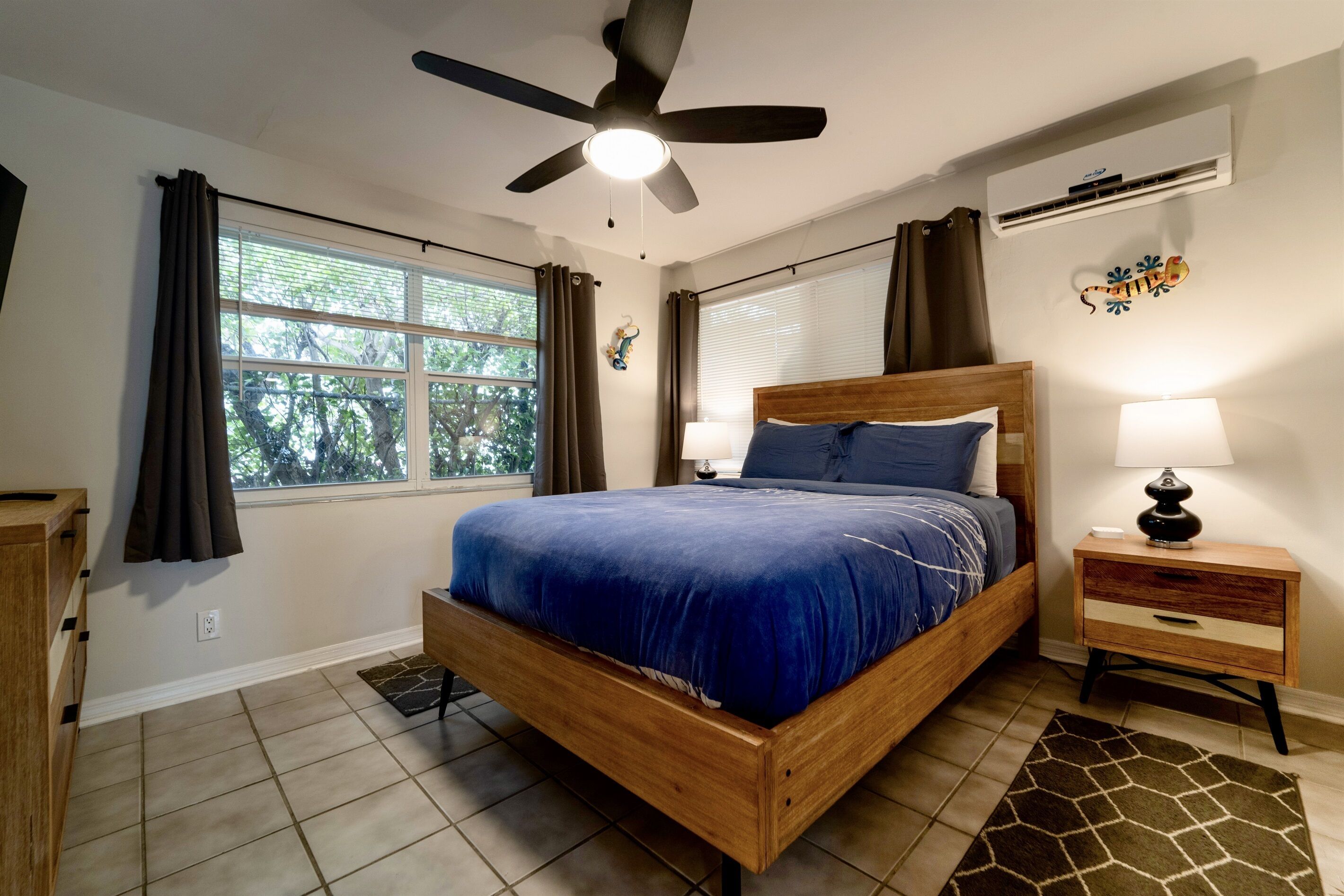 A bedroom with a wooden bed, a ceiling fan, a nightstand with a lamp, and a window with curtains.