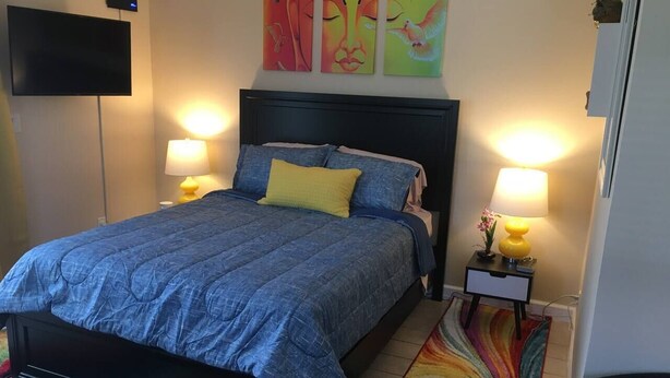Ein Schlafzimmer mit einem Bett, zwei Nachttischlampen, einem Fernseher und farbenfroher Wandkunst.
