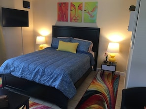 A bedroom with a bed, two bedside lamps, a TV, and colorful artwork on the wall.