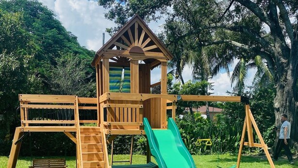 Uma estrutura de recreio de madeira com escorrega, escada de escalada e uma pequena casinha.