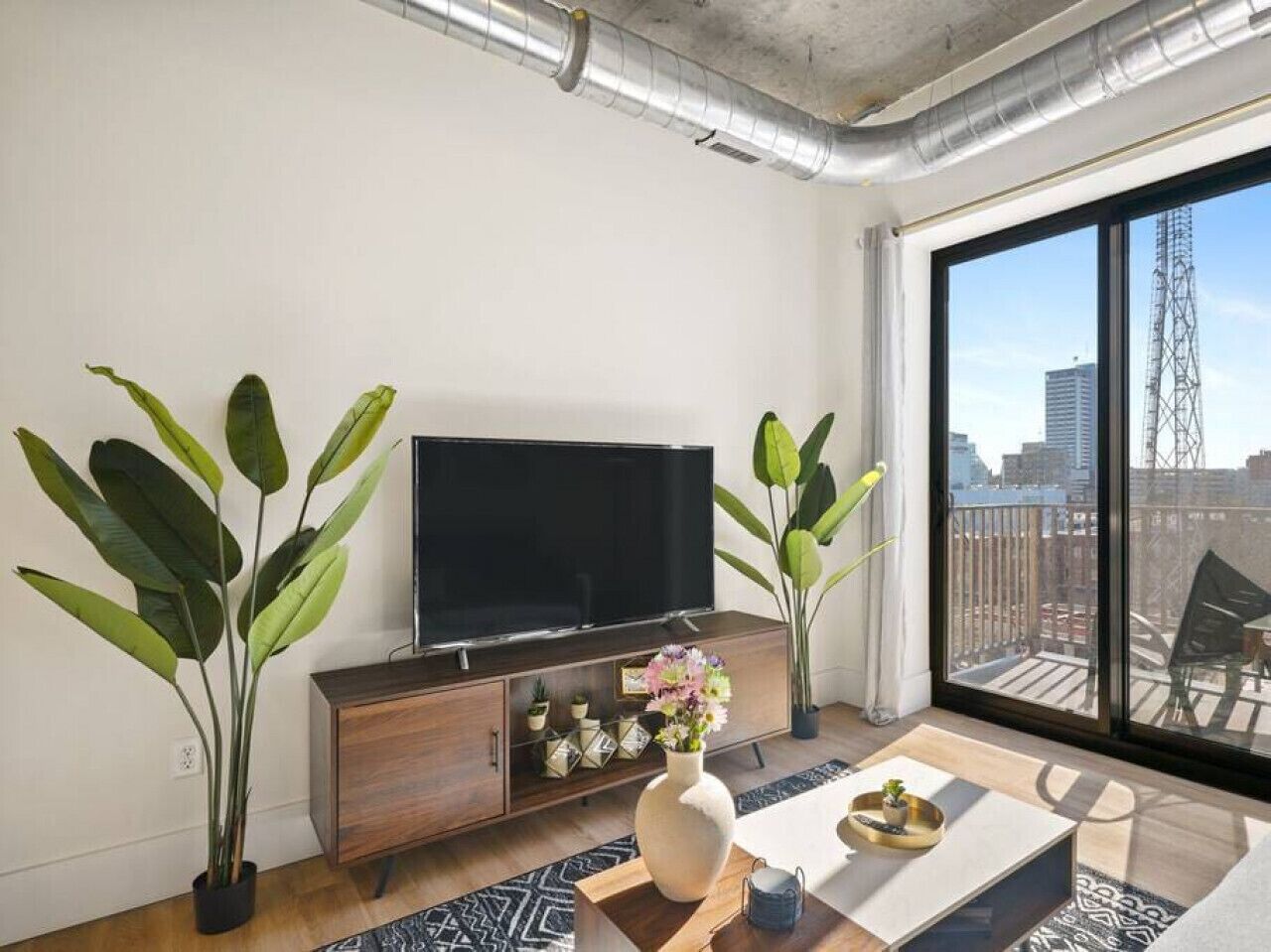 A modern living room with a large flat-screen TV, wooden cabinet, and two large potted plants.