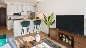A modern living room with a flat-screen TV, two green bar stools, a coffee table with a vase, and a kitchen area in the background. - Clover Haven | High Rise | Down Town South Bend ND (South Bend)