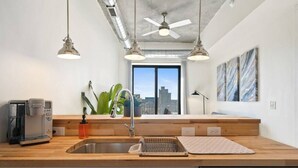 A modern kitchen with a wooden countertop, stainless steel sink, and a dishwasher. There are pendant lights, a coffee maker, and a plant on the counter. - Clover Haven | High Rise | Down Town South Bend ND (South Bend)