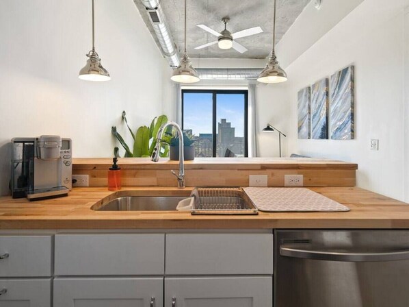 A modern kitchen with a wooden countertop, stainless steel sink, and a dishwasher. There are pendant lights, a coffee maker, and a plant on the counter.