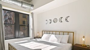 A bedroom with a bed, a nightstand, a window, and a wall with moon phases. - Clover Haven | High Rise | Down Town South Bend ND (South Bend)