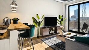 A modern living space with a kitchen, dining area, and seating area. It features a large window with a city view, a flat-screen TV, and a ceiling fan. - Clover Haven | High Rise | Down Town South Bend ND (South Bend)