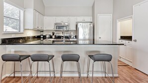 Een moderne keuken met witte kastjes, roestvrijstalen apparatuur en een zwart aanrecht. Drie barstoelen en een raam met zonwering.