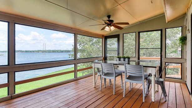 Een overdekt buitengebied met een houten terras, een tafel met stoelen en grote ramen met uitzicht op het water en groen.