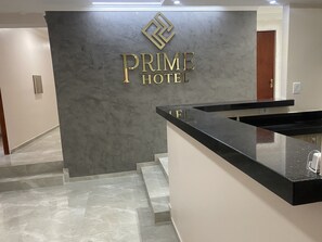 Zona de recepción de un hotel con piso de mármol, un mostrador de recepción y una pared con el nombre del hotel en letras doradas.