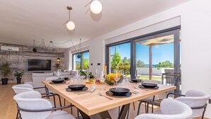 A modern dining area with a wooden table, grey upholstered chairs, and a large window offering a view of the outdoors. - Gorgeous home in Sibenik (Sibenik)
