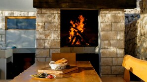A stone fireplace with a fire, a wooden table with bread and olives, and a stone wall.