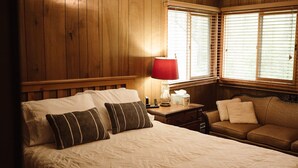 A bedroom with a wooden paneled wall, a bed with two pillows, a nightstand with a lamp, and a window with blinds.