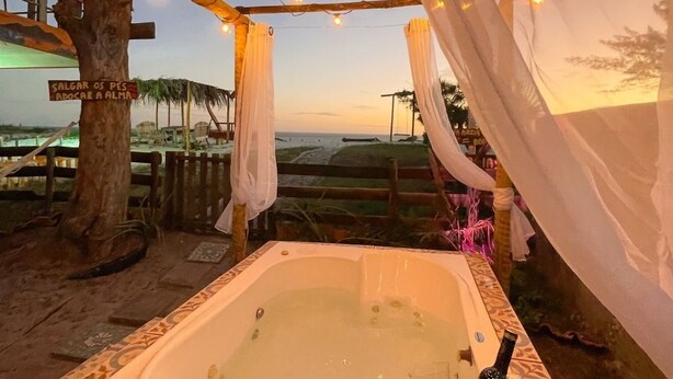 A hot tub under a wooden pergola with string lights, surrounded by a rustic setting with a tree and a view of the landscape. - Pé na areia. (Arraial do Cabo)
