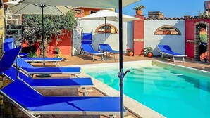 Zona de piscina con sillas de descanso azules y sombrillas, rodeada por un edificio rojo y blanco con ventanas arqueadas y plantas en macetas.