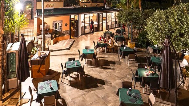 Un patio al aire libre con mesas y sillas, un edificio con techo de tejas y follaje en el fondo.