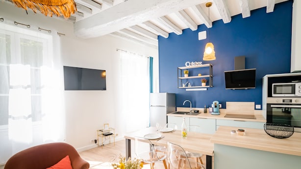 Eine moderne Küche mit blauer Akzentwand, ein Essbereich mit Holztisch und -stühlen sowie eine gemütliche Sitzecke mit einem roten Sessel und einem Blumenvasen.
