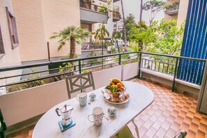A balcony with a table set for tea, surrounded by greenery and a view of neighboring buildings.