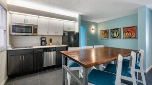 Ett modernt kök med vita skåp, en mikrovågsugn i rostfritt stål och ett svart kylskåp. Det finns ett matbord i trä med blå stolar och tre abstrakta tavlor på väggen.