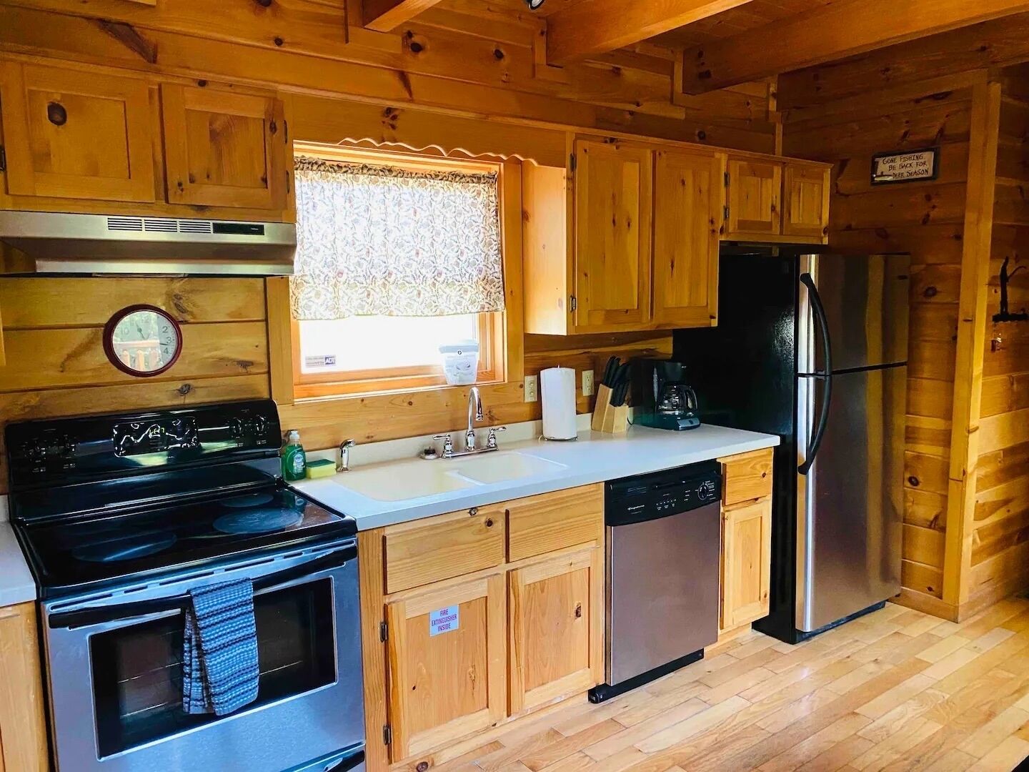 A compact kitchen with wooden cabinets, a stainless steel refrigerator, a dishwasher, a stove, and a clock on the wall.