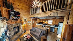 Ein gemütliches Blockhaus-Interieur mit Kamin aus Stein, einem Wohnbereich mit Sofa und Couchtisch, einer Küche mit Holzschränken und einem Balkon mit Holzgeländer.
