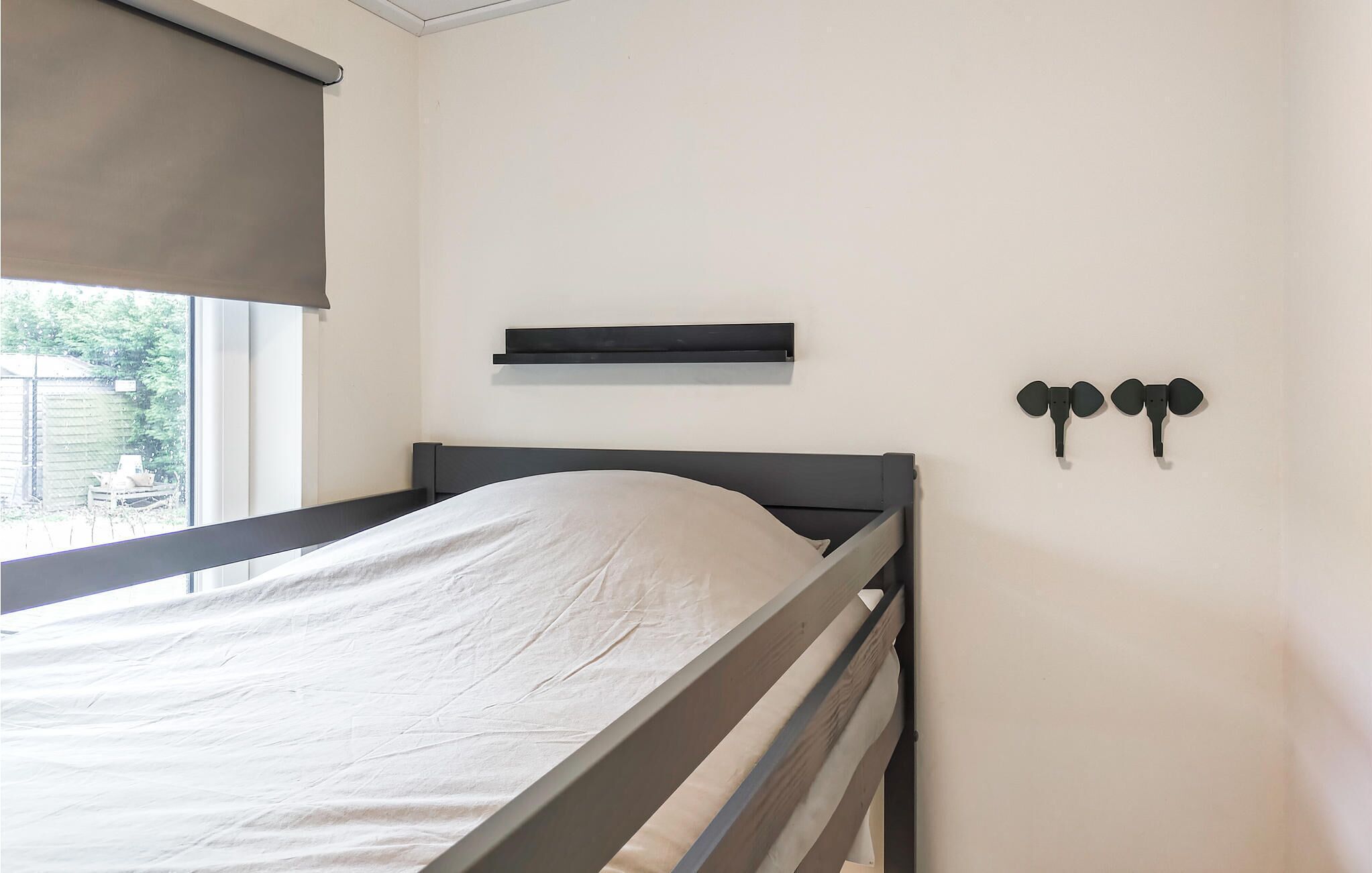 A single bed with a headboard, a window with a blind, a shelf above the bed, and two wall-mounted hooks.