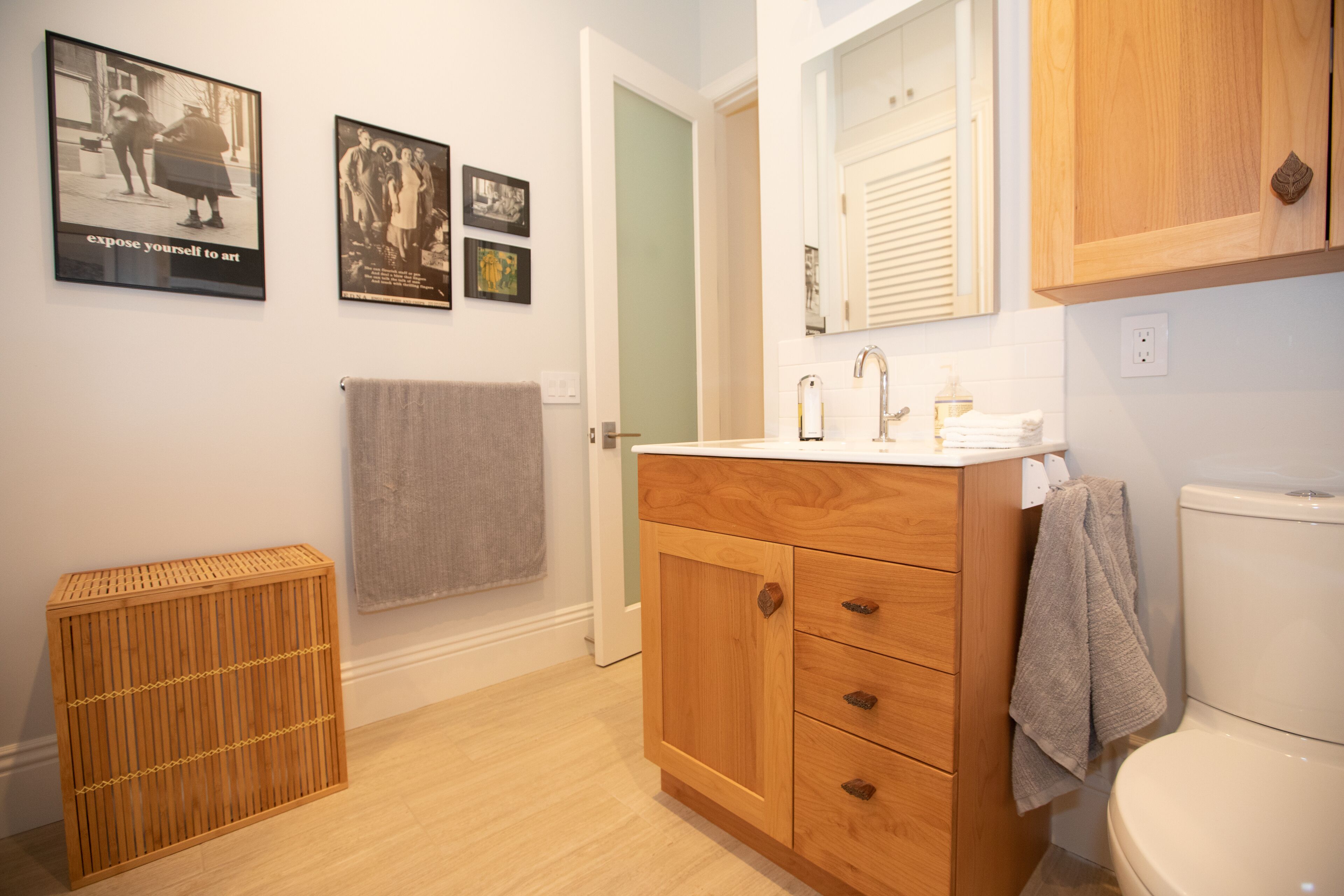 A bathroom with wooden cabinets, a white sink, and a toilet. There are two framed pictures on the wall and a wooden stool.