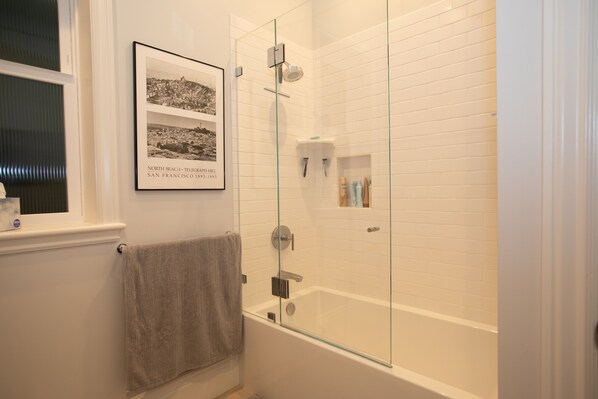 A modern bathroom with a glass-enclosed shower, a white bathtub, and a framed historical photograph on the wall.