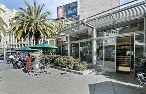 An outdoor café with tables and chairs, palm trees, and a building with glass doors and a sign.