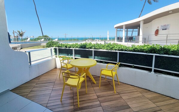 A balcony with a wooden floor, yellow chairs, and a round table. - Frente Mar com Terraço Privativo e Pet Friendly (Barra de São Miguel)
