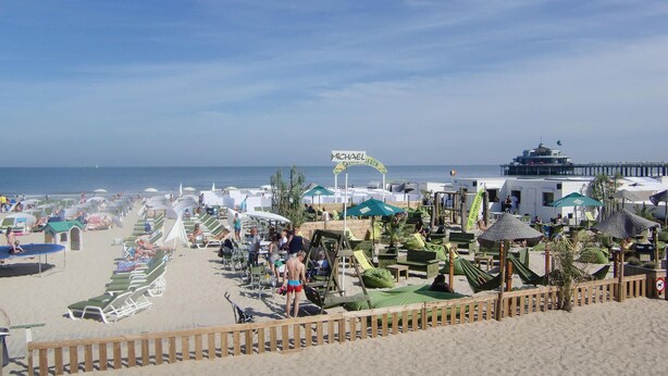 Un complexe hôtelier en bord de mer, avec des rangées de chaises longues, une clôture en bois et un panneau indiquant « Michael’s Beach Club ».