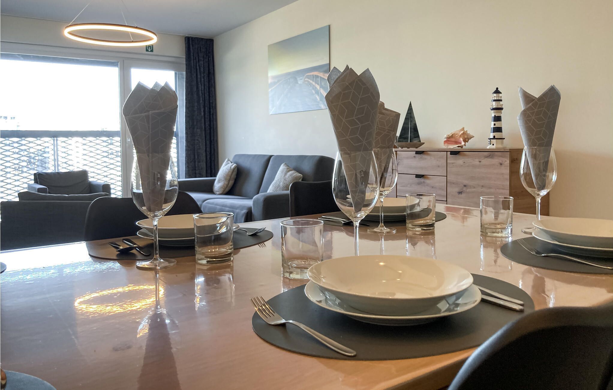 A dining table set with plates, glasses, and cutlery in a modern room with a sofa and decorative items in the background.