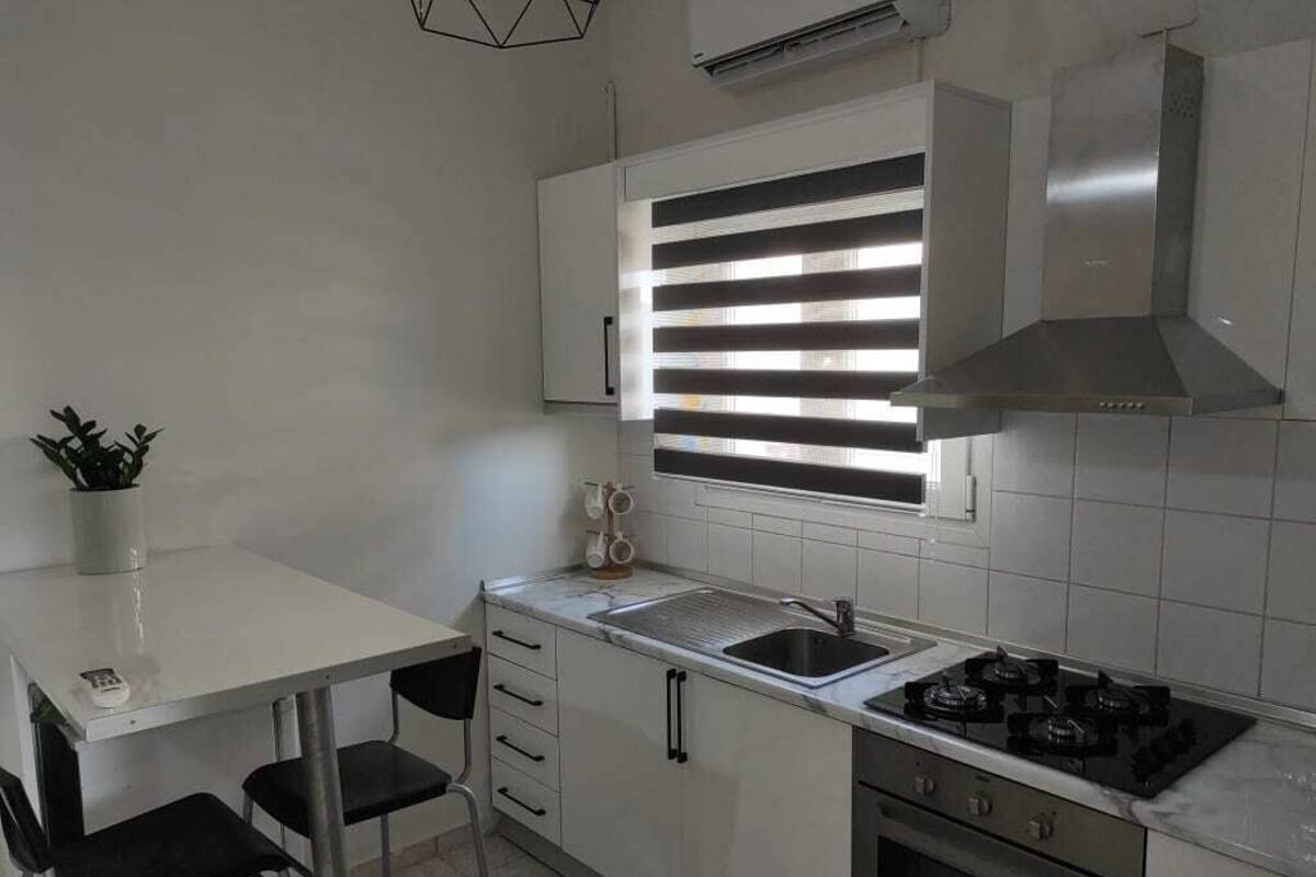 Une cuisine avec un plan de travail blanc, un évier, une cuisinière et une fenêtre avec des stores.