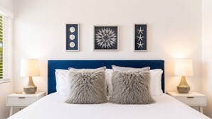 A bed with two fluffy pillows, a blue headboard, and three framed artworks on the wall.