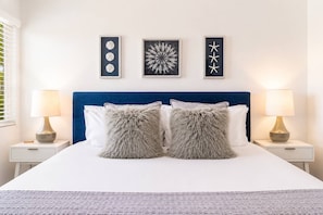 A bed with two fluffy pillows, a blue headboard, and three framed artworks on the wall.