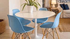 A dining area with a round table, blue chairs, a potted plant, and a patterned wallpaper background.