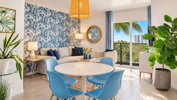 A modern living room with a sofa, a round dining table with chairs, a large window with a view, and a tropical wallpaper.