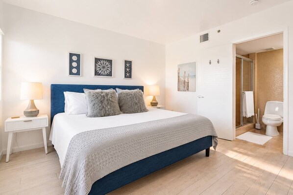 A bedroom with a bed, two bedside tables, a nightstand, a door, and a bathroom visible through an open door.