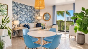 A modern living room with a sofa, a round dining table with chairs, a large window with a view, and a tropical wallpaper. - Walk to Beach- South of Fifth Condo | Free Parking (Miami Beach)