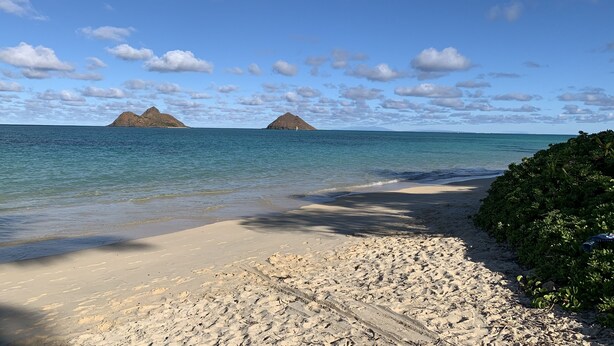 Une plage à l’eau d’un bleu limpide, bordée d’un rivage sablonneux, avec des montagnes qui se dessinent à l’horizon.