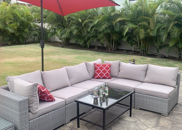 A sectional sofa with light cushions and red patterned pillows, a glass-top coffee table, and a red umbrella. - Bright, upgraded house, A/C, near the ocean! CONTACT FOR RATES & AVAILABILITY! (Kailua)