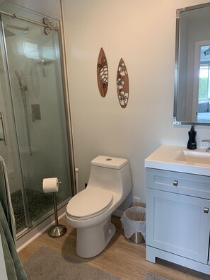 A bathroom with a glass shower enclosure, a toilet, a sink with a mirror, and decorative wall art. - Bright, upgraded house, A/C, near the ocean! CONTACT FOR RATES & AVAILABILITY! (Kailua)