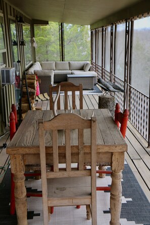 A wooden table with chairs, a sofa, and a view of trees through large windows on a covered deck.