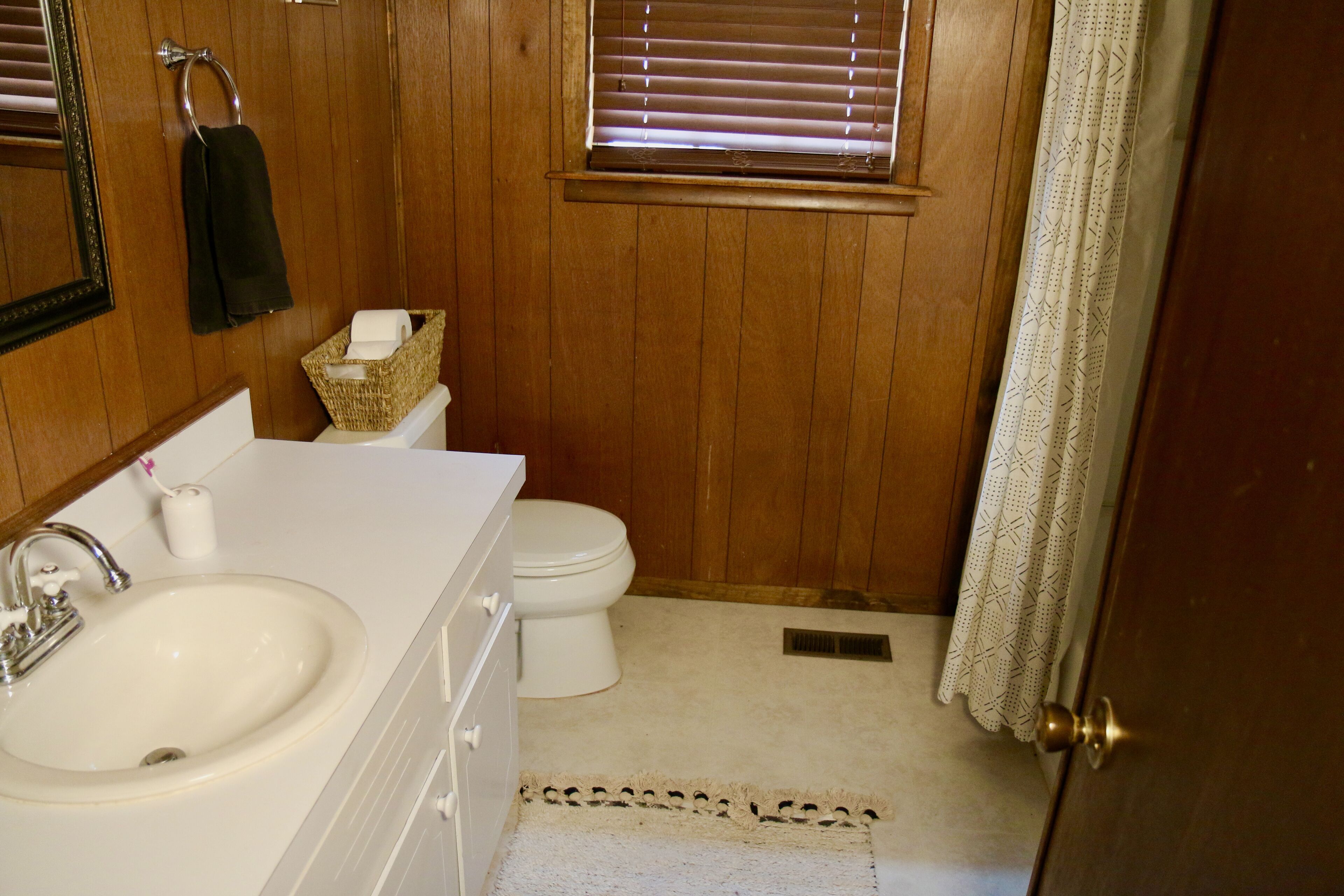 Une salle de bain avec un mur en bois, un lavabo blanc, des toilettes et une fenêtre avec des stores.