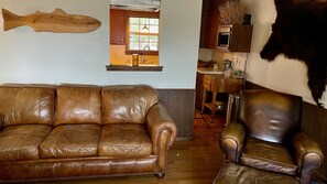A brown leather sofa and armchair in a living room with a wooden fish mounted on the wall, a window with a view, and a kitchen area in the background.