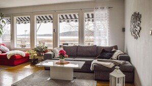 A living room with a grey sofa, a red armchair, and a white coffee table. There are large windows with striped blinds, a view of a body of water, and a metal wall art piece. - Pet friendly home in Trosa with sauna (Trosa)