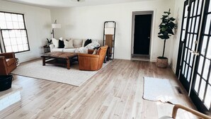 Ein geräumiges Wohnzimmer mit Holzboden, einer Couch, einem Couchtisch und einem Beistelltisch. Es gibt ein großes Fenster mit Jalousien, eine Tür und einen Blumentopf.